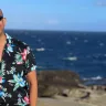 A man in a Hawaiian shirt standing on a rocky beach with the ocean in the background